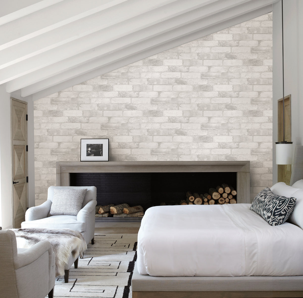 Modern bedroom featuring York Wallcoverings’ Reclaimed Bricks Wallpaper as a white brick accent wall, exposed ceiling beams, a bed with white bedding, a cozy armchair, fireplace with stacked firewood, and minimalist black-and-white decor.