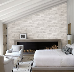 Modern bedroom featuring York Wallcoverings’ Reclaimed Bricks Wallpaper as a white brick accent wall, exposed ceiling beams, a bed with white bedding, a cozy armchair, fireplace with stacked firewood, and minimalist black-and-white decor.