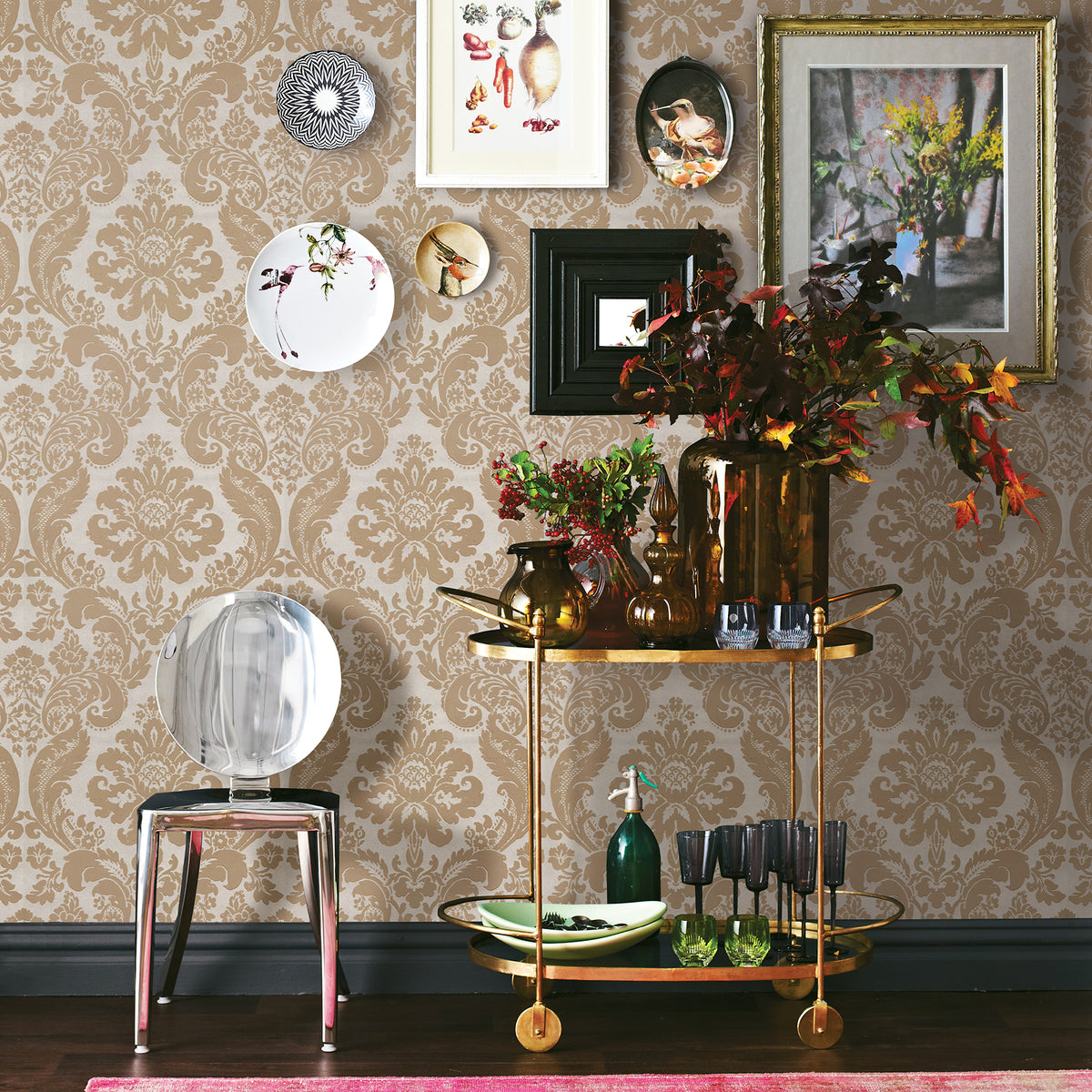 A gold bar cart stocked with glassware, bottles, and autumn-inspired floral arrangements stands beside a clear chair, set against York Wallcoverings’ Shadow Flocked Damask Wallpaper adorned with decorative plates and framed artwork.
