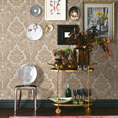 A gold bar cart stocked with glassware, bottles, and autumn-inspired floral arrangements stands beside a clear chair, set against York Wallcoverings’ Shadow Flocked Damask Wallpaper adorned with decorative plates and framed artwork.