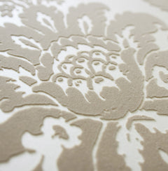 Close-up of the Shadow Flocked Damask Wallpaper by York Wallcoverings, featuring a beige, raised damask floral pattern with detailed petals and leaves on a textured surface set against a smooth, off-white background.