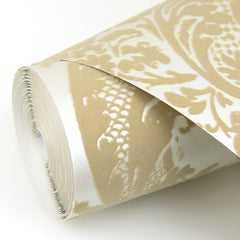 A close-up of the York Wallcoverings Shadow Flocked Damask Wallpaper, featuring an ornate beige and white floral damask pattern, rolled and displayed on a white surface.