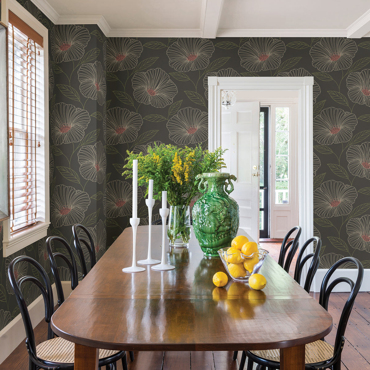 A dining room featuring a wooden table, black chairs, and sunlight streaming through wooden blinds. The walls are adorned with Mythic Wallpaper by York Wallcoverings in a green floral pattern. The table is decorated with candles, a glass vase filled with yellow flowers, a large green vase, and a bowl of lemons.