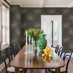 A dining room featuring a wooden table, black chairs, and sunlight streaming through wooden blinds. The walls are adorned with Mythic Wallpaper by York Wallcoverings in a green floral pattern. The table is decorated with candles, a glass vase filled with yellow flowers, a large green vase, and a bowl of lemons.