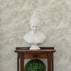 A white marble bust of a woman rests on a wooden table with a small green plant underneath, set against Reyne Marble Wallpaper by York Wallcoverings in a light gray marbled finish.