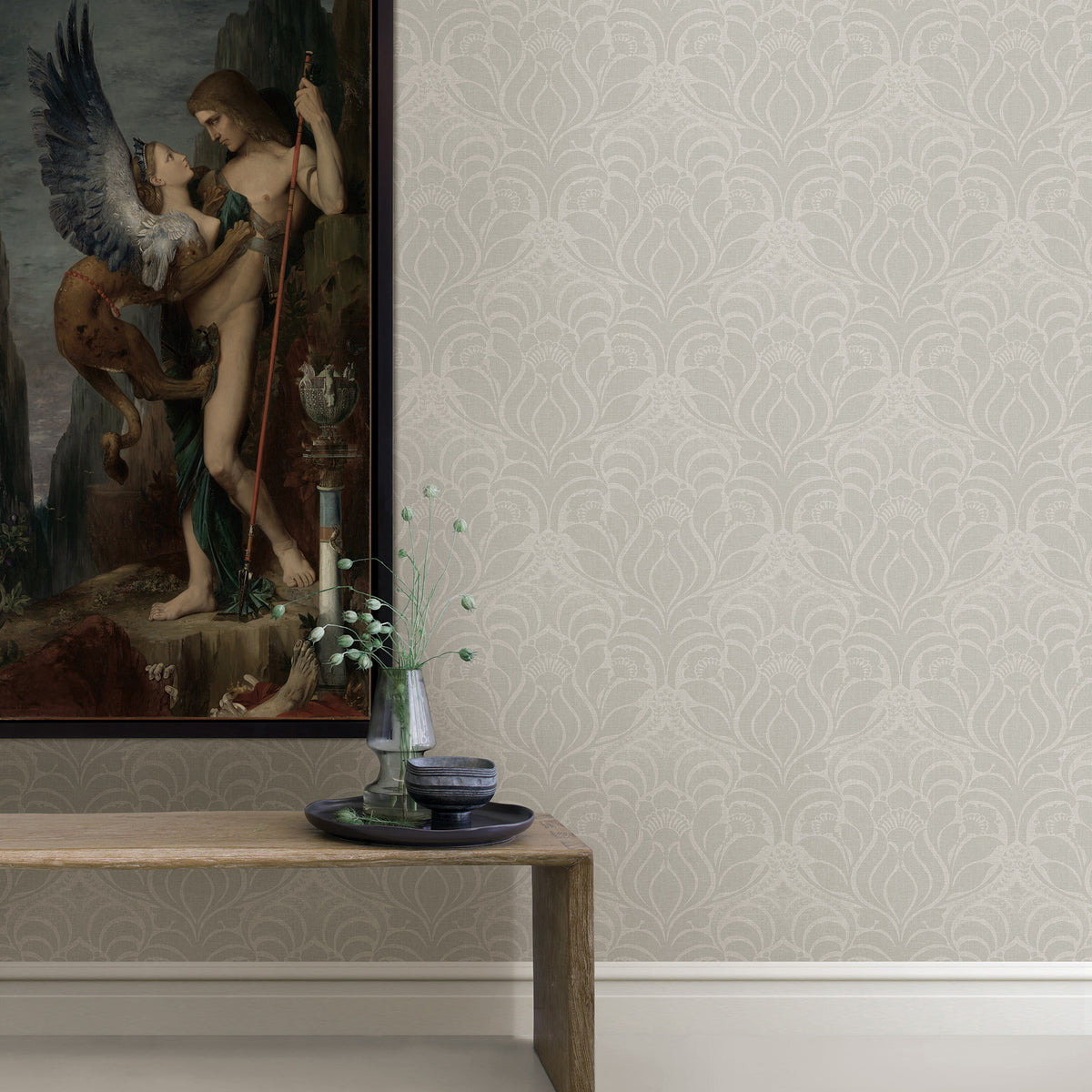 A classical painting hangs on a wall adorned with Sandor Damask Wallpaper by York Wallcoverings above a wooden table holding a glass vase with greenery, a bowl, and a plate.