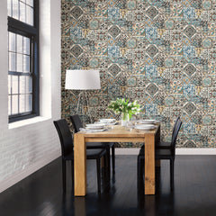 A dining area with a wooden table, four black chairs, a white floor lamp, and a vase of flowers. The wall features York Wallcoverings’ Estrada Marrakesh Tiles Wallpaper in colorful, intricate patterns; large windows let natural light pour onto dark wood flooring.