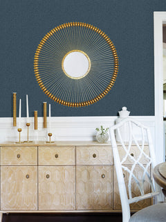 A modern room featuring York Wallcoverings’ Hilbert Geometric Wallpaper in dark blue, a gold sunburst mirror, a light wood sideboard, gold candleholders, and a white chair with a geometric backrest.
