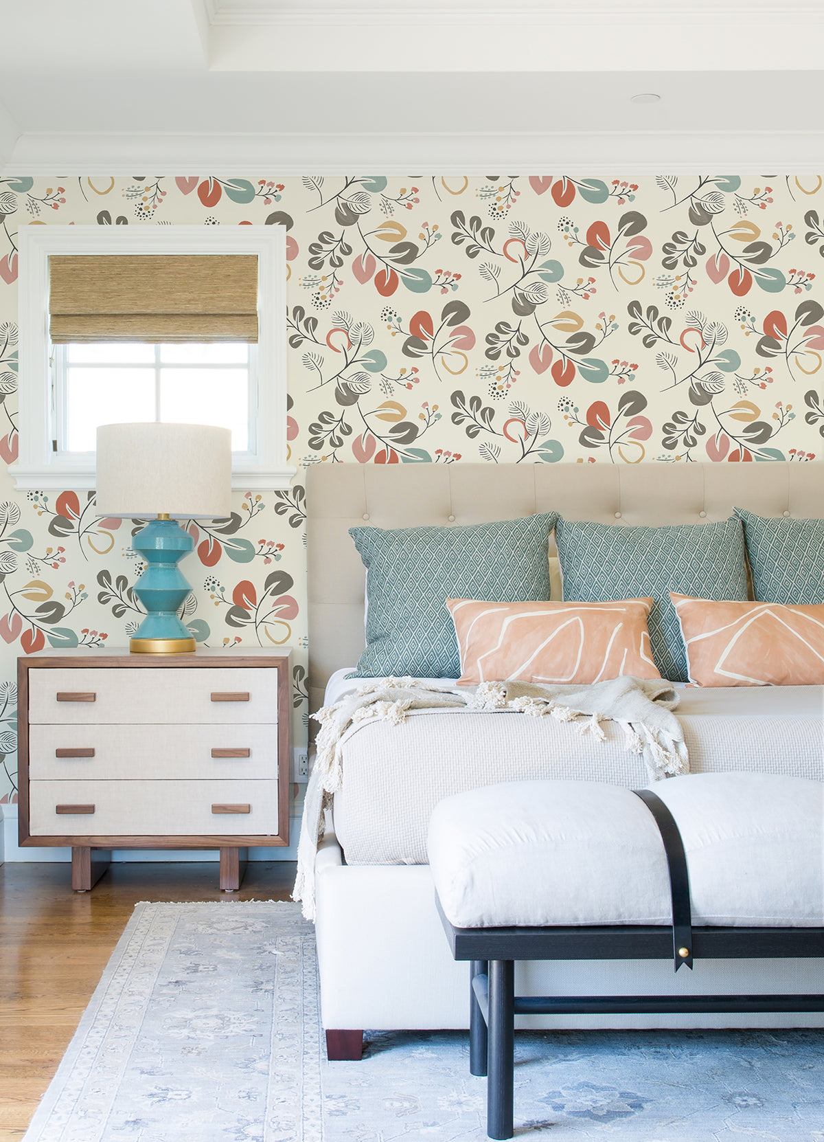 Bright bedroom featuring York Wallcoverings’ Jonah Leaf Trail Wallpaper, a bed with blue and peach pillows, a light bench, a small dresser with a blue lamp, a window with a bamboo shade, and a blue rug on hardwood floors.