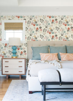 Bright bedroom featuring York Wallcoverings’ Jonah Leaf Trail Wallpaper, a bed with blue and peach pillows, a light bench, a small dresser with a blue lamp, a window with a bamboo shade, and a blue rug on hardwood floors.