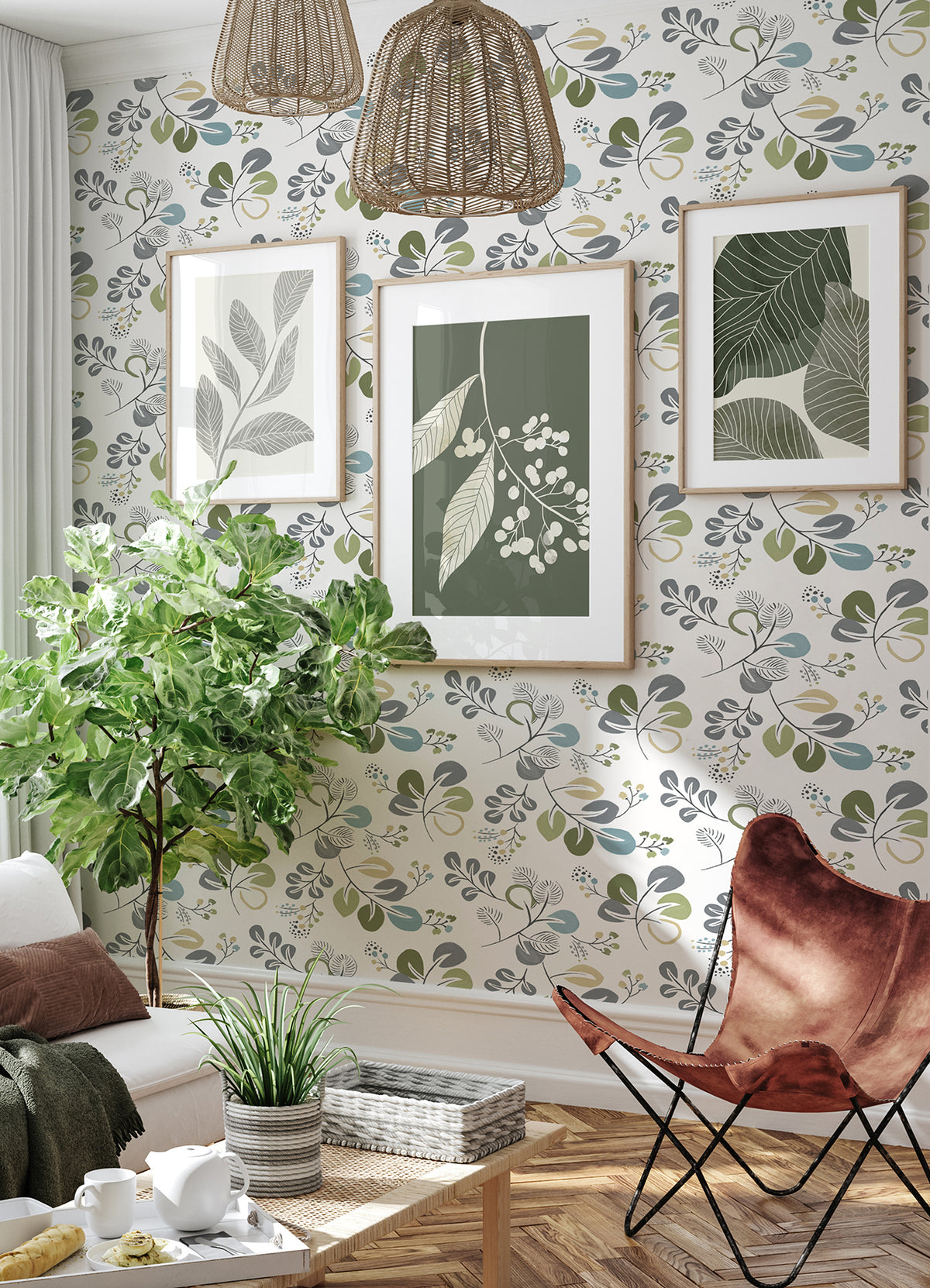 A cozy living room featuring York Wallcoverings’ Jonah Leaf Trail Wallpaper, three framed botanical prints on the wall, a large leafy plant, wicker lamps, a brown butterfly chair, and a white sofa accented with a green pillow and blanket.