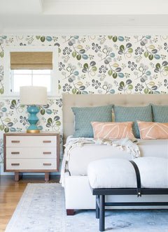 Bright bedroom featuring Jonah Leaf Trail Wallpaper by York Wallcoverings, bed with blue and peach pillows, white tufted bench, light wood nightstand, blue lamp, and a window dressed with a bamboo Roman shade. A light rug and hardwood floor complete the space.
