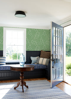 A cozy room featuring York Wallcoverings’ Elin Berry Botanical Wallpaper in green, a built-in bench with striped and patterned pillows, a round wooden table topped with a glass vase, a striped rug, and an open door letting in sunlight.