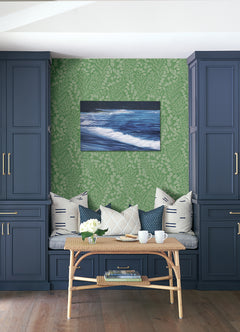 Blue built-in cabinets frame a cozy bench with patterned pillows, a wicker table with mugs, a vase of flowers, and Elin Berry Botanical Wallpaper by York Wallcoverings in lush green leafy patterns. An ocean wave painting hangs above the bench.