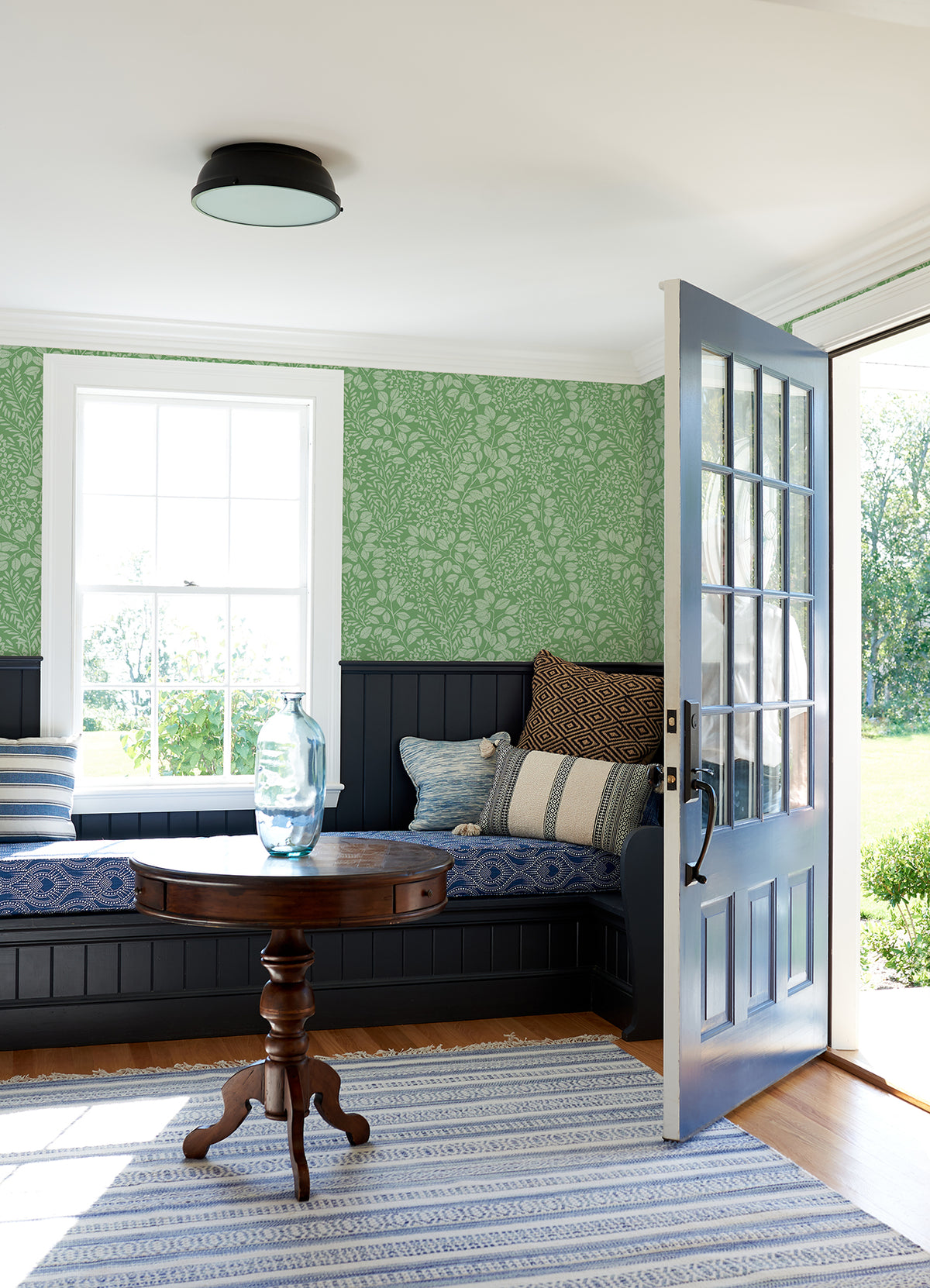 A cozy room with a blue door open to the outside features a round wooden table topped with a glass vase, built-in bench seating with cushions, York Wallcoverings’ Elin Berry Botanical Wallpaper in green on the walls, and a striped rug on the floor.