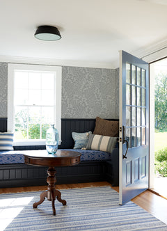 A cozy room featuring a blue-patterned bench, various pillows, a round wooden table with a glass vase, an open blue door, striped rug, and a bright window framed by Elin Berry Botanical Wallpaper from York Wallcoverings.
