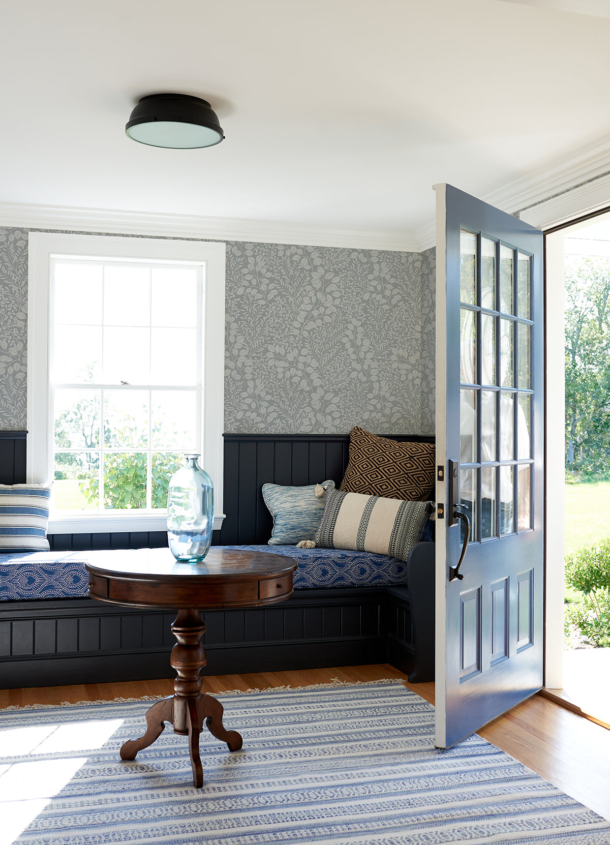 A sunlit room features a cozy corner bench adorned with patterned blue cushions, a round wooden table topped with a glass vase, and walls decorated in Elin Berry Botanical Wallpaper by York Wallcoverings. An open blue door leads outside, and a striped rug lies over the wooden floor.