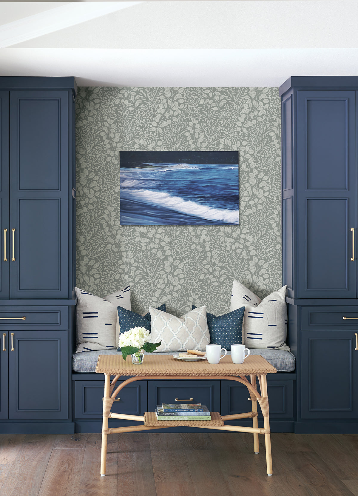 A cozy bench with patterned pillows sits between blue cabinets. In front, a rattan table holds white mugs, a teapot, and flowers. On the wall, ocean artwork is displayed against Elin Berry Botanical Wallpaper by York Wallcoverings.