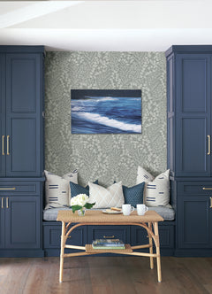 A cozy bench with patterned pillows sits between blue cabinets. In front, a rattan table holds white mugs, a teapot, and flowers. On the wall, ocean artwork is displayed against Elin Berry Botanical Wallpaper by York Wallcoverings.