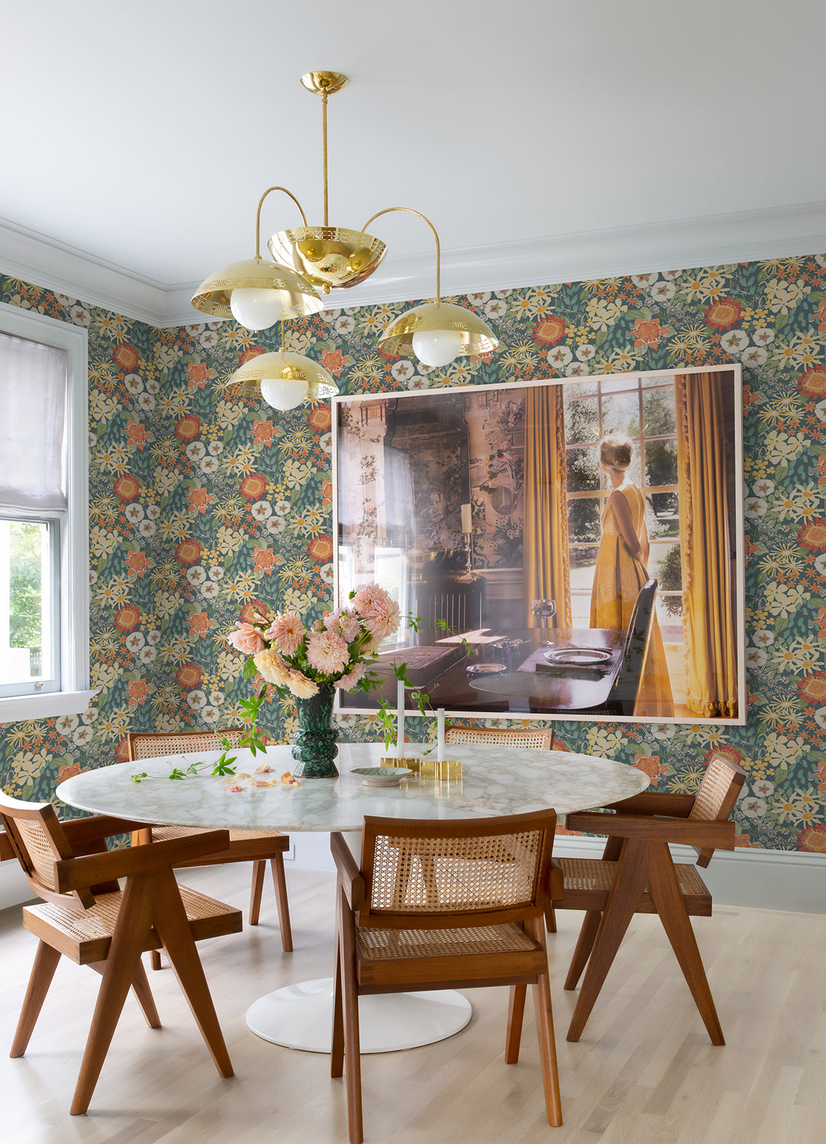 A dining area featuring York Wallcoverings’ Karina Wildflower Garden Wallpaper, a round white table with wooden chairs, a gold chandelier, a vase of pink flowers, and a large photo of a woman in a yellow dress hanging on the wall beside yellow curtains.