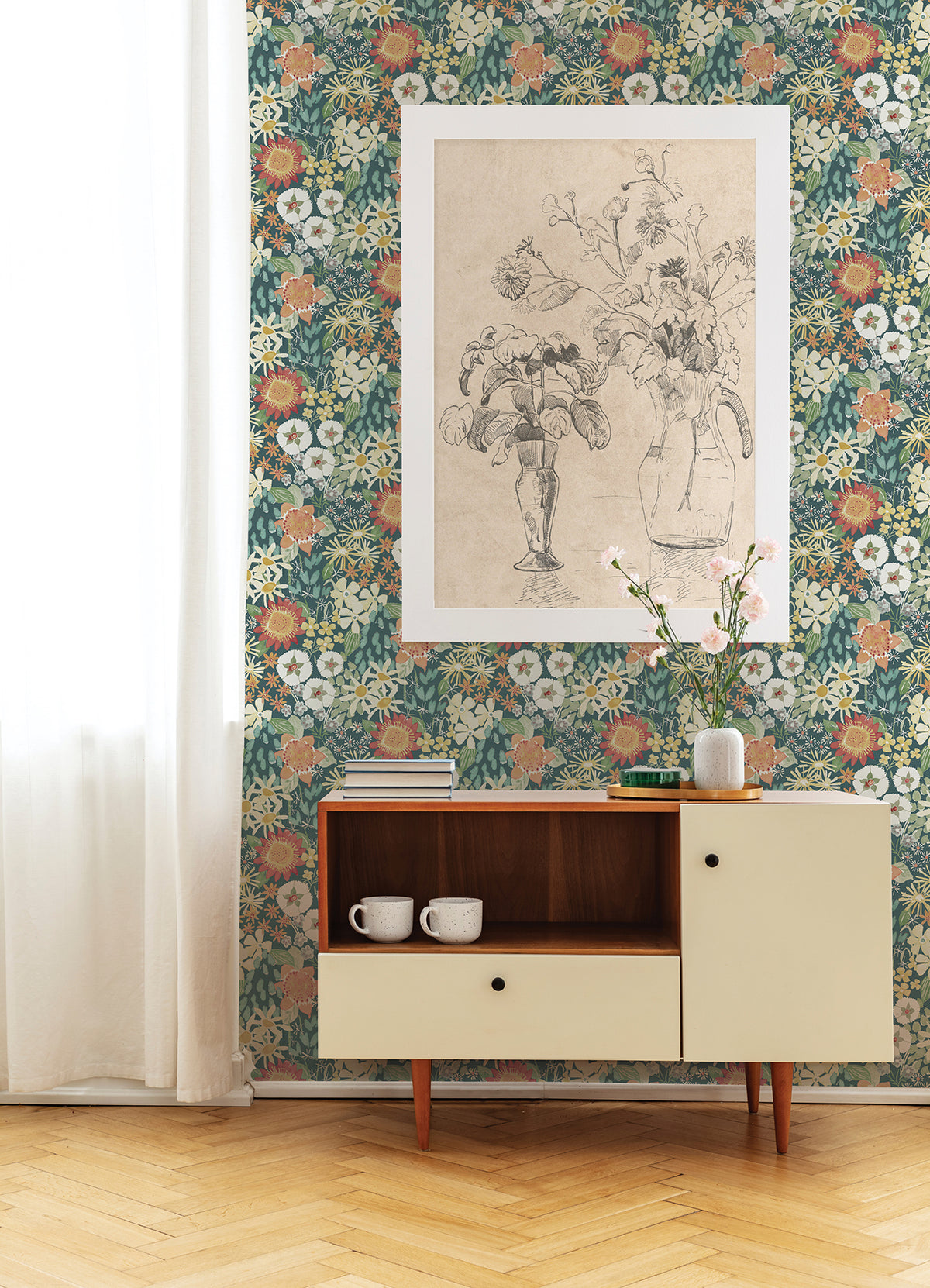 A sideboard with two mugs sits against a wall adorned in Karina Wildflower Garden Wallpaper by York Wallcoverings. Above is a framed black-and-white flower sketch, while a vase with pink flowers rests on the sideboard; white curtains hang to the left.