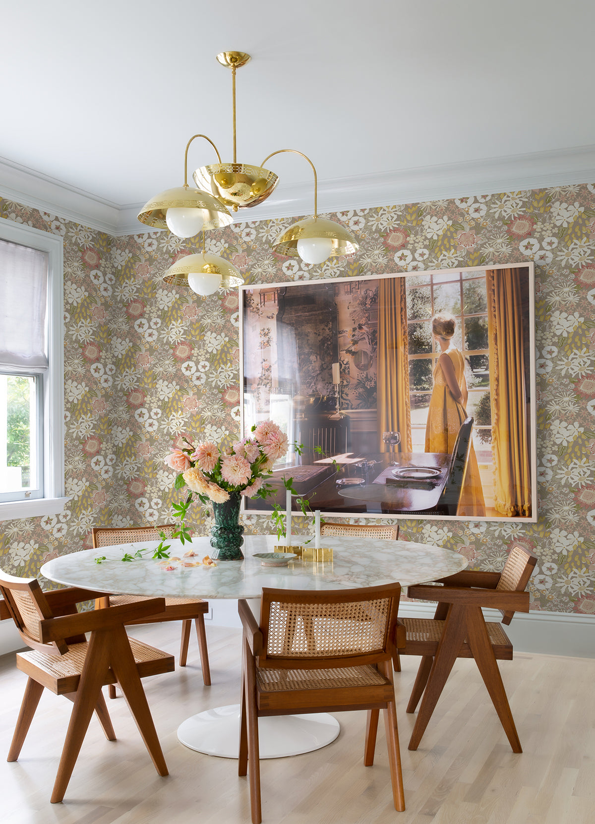 Dining area featuring a round marble table, wooden chairs, a vase of flowers, a gold chandelier, and a large framed photo of a woman standing by a sunny window with yellow curtains. The walls are adorned with Karina Wildflower Garden Wallpaper by York Wallcoverings.