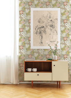 A mid-century modern cabinet with two white mugs stands against a wall adorned with Karina Wildflower Garden Wallpaper by York Wallcoverings, featuring a large botanical sketch above. A single flower in a vase sits atop the cabinet.
