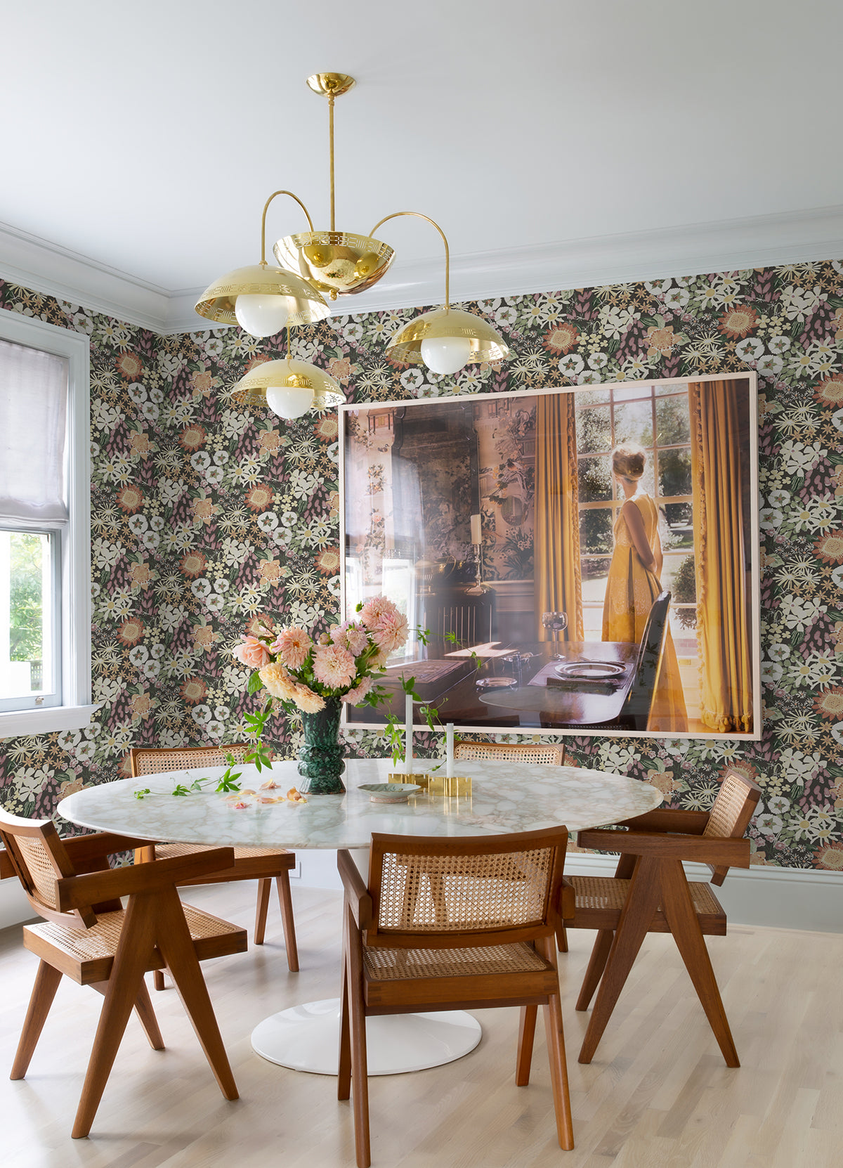 A dining area featuring a round white table, wooden chairs, a vase of pink flowers, a gold chandelier, and a framed photo of a woman in a yellow dress looking out at yellow curtains, with the walls adorned in York Wallcoverings’ Karina Wildflower Garden Wallpaper.