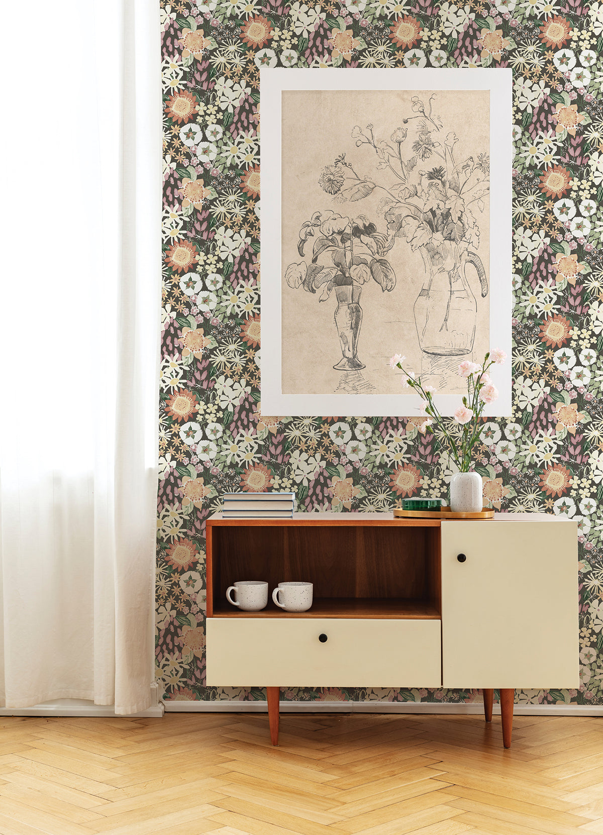 A mid-century cabinet sits against a wall adorned with Karina Wildflower Garden Wallpaper by York Wallcoverings. On the cabinet are two white mugs and a vase with pink flowers, while above hangs a framed floral sketch. Sunlight streams through a sheer white curtain on the left.