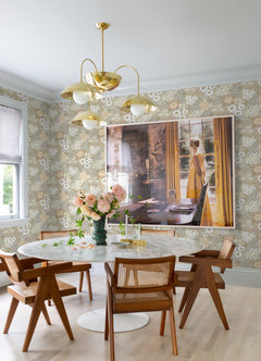 A bright dining room featuring a round white table, four wooden chairs, a vase of flowers, a gold chandelier, and large art of a woman in a yellow dress by a window with yellow curtains, with the walls adorned in Karina Wildflower Garden Wallpaper by York Wallcoverings.