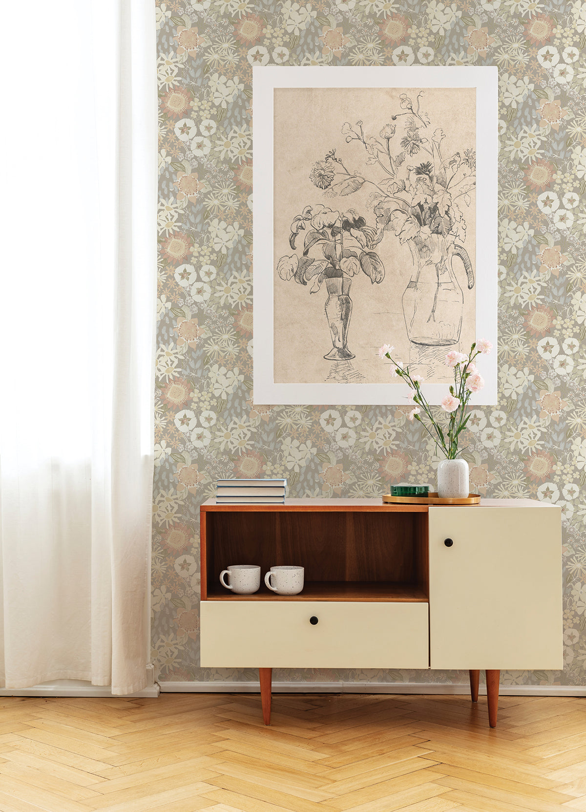 Against a wall adorned with York Wallcoverings’ Karina Wildflower Garden Wallpaper, a mid-century cabinet showcases two white mugs, a vase of flowers, and several books. A large botanical sketch is displayed nearby while soft light streams in through a sheer curtain on the left.