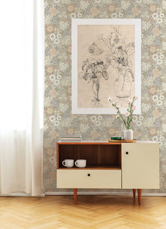 Against a wall adorned with York Wallcoverings’ Karina Wildflower Garden Wallpaper, a mid-century cabinet showcases two white mugs, a vase of flowers, and several books. A large botanical sketch is displayed nearby while soft light streams in through a sheer curtain on the left.