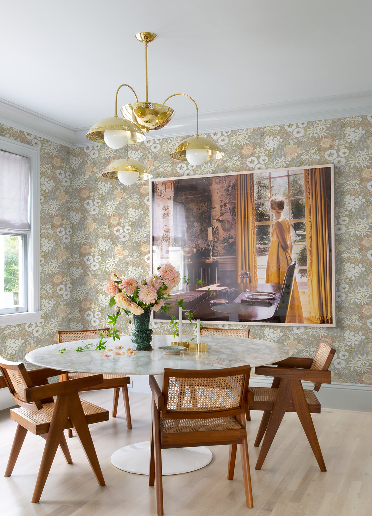 A dining room featuring a round marble table and wooden chairs, adorned with York Wallcoverings’ Karina Wildflower Garden Wallpaper, a gold chandelier, and a large framed photo of a woman in a yellow dress by a window. Pink flowers and glassware are arranged on the table.