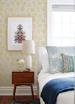 A cozy bedroom corner featuring a mid-century nightstand, white lamp, small vase with flowers, framed floral art, a bed decorated with blue and white pillows and a blue throw, all set against York Wallcoverings’ Inge Floral Block Print Wallpaper.