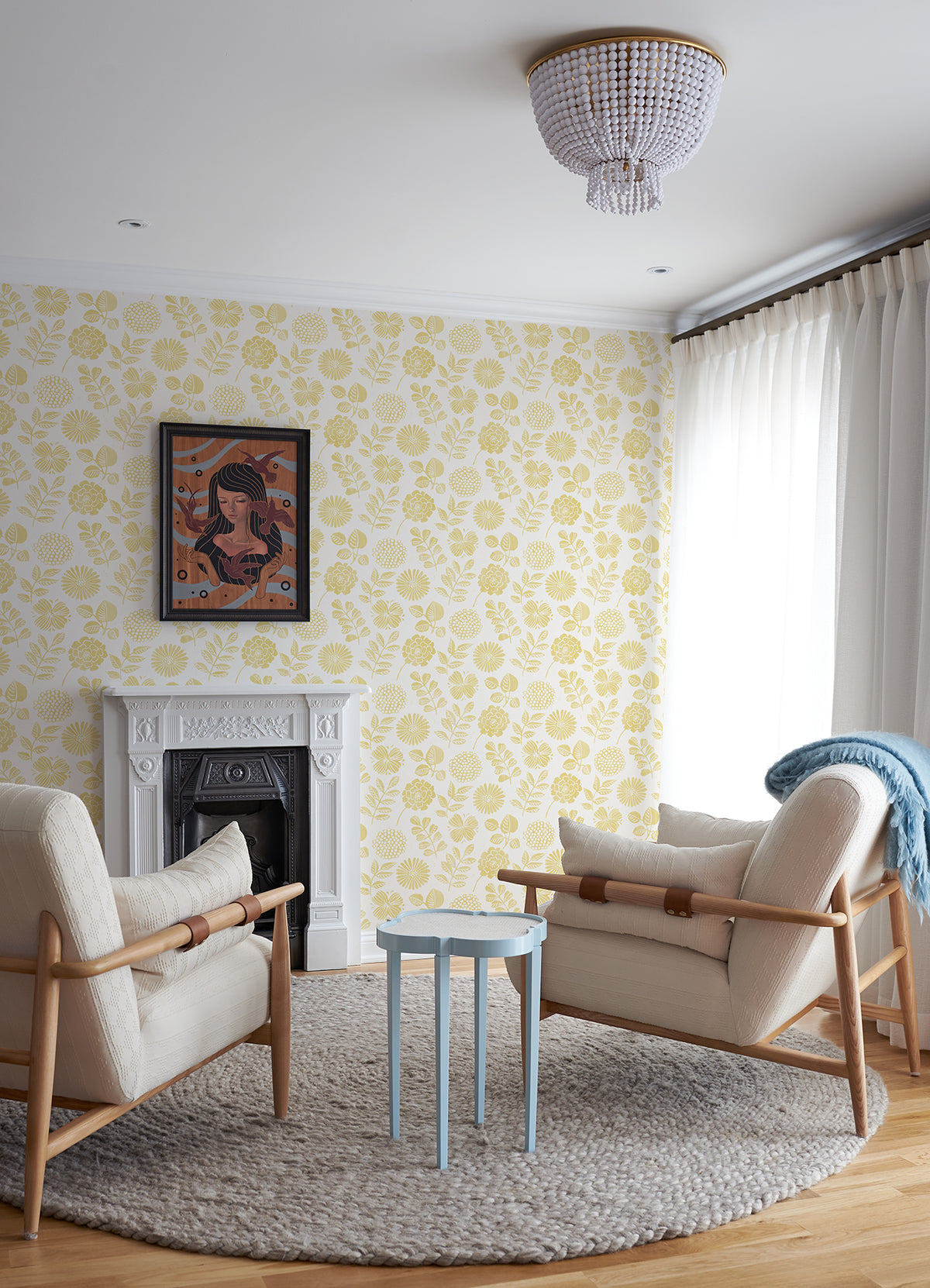 Two armchairs face a white fireplace in a cozy living room with York Wallcoverings’ Inge Floral Block Print Wallpaper, a round gray rug, a light blue side table, curtains, and a ceiling light. A framed artwork hangs above the fireplace.