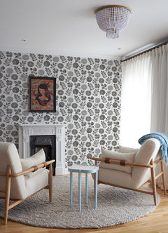 Modern living room with two cream armchairs, a small blue table, and a round rug. The Inge Floral Block Print Wallpaper by York Wallcoverings adds charm behind a white fireplace, while framed art and sheer curtains complete the cozy, stylish atmosphere.