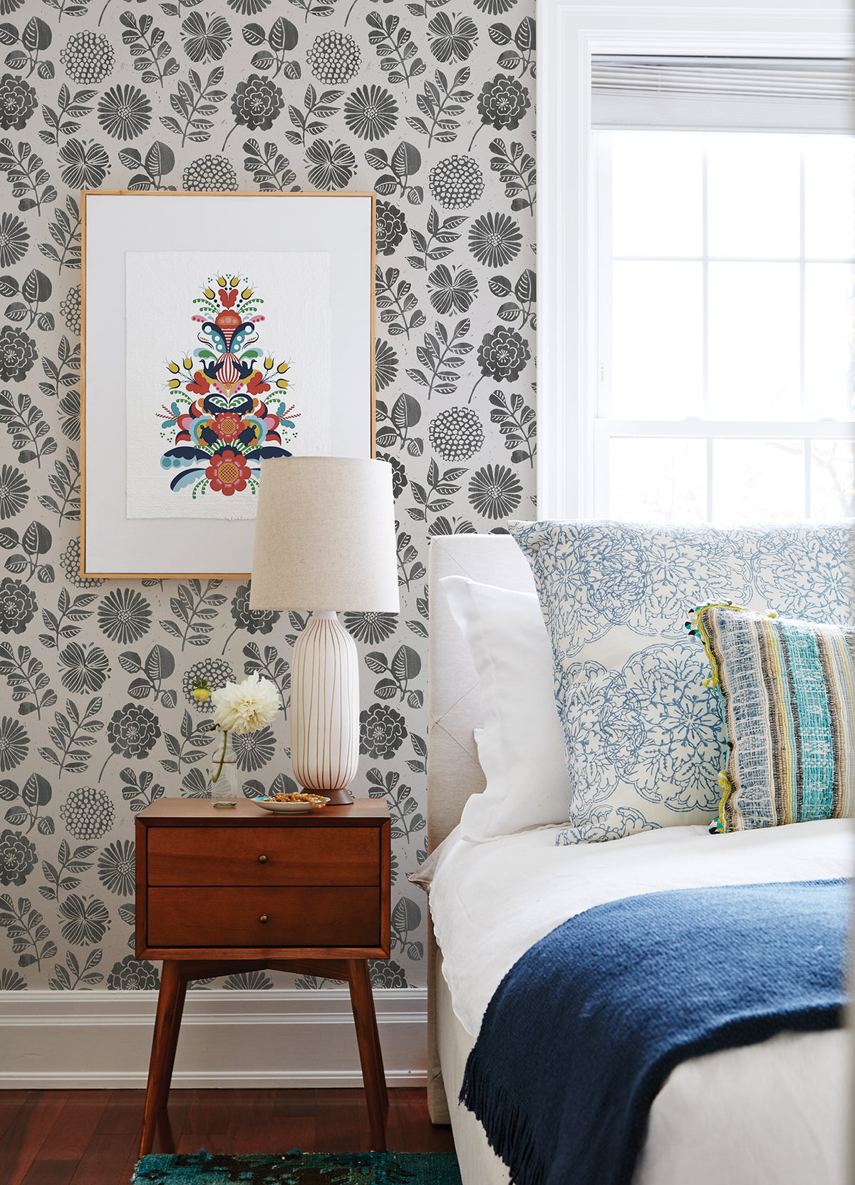 A bedside table with a lamp, small vase, and books sits next to a bed with blue and white bedding, set against a wall covered in Inge Floral Block Print Wallpaper by York Wallcoverings. A colorful framed artwork hangs above the table while sunlight streams through a window.