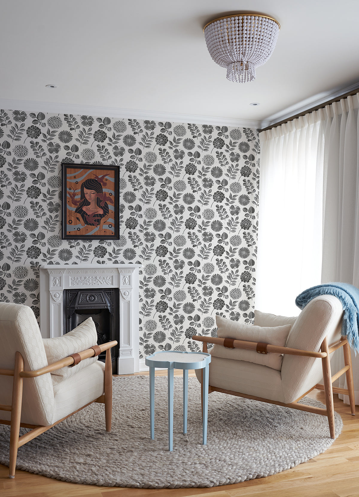 Modern living room with two cream armchairs, a small blue table, a round gray rug, white fireplace, sheer white curtains, and York Wallcoverings’ Inge Floral Block Print Wallpaper in black and white. A framed artwork hangs above the fireplace.