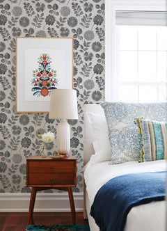 A bedside table with a lamp, small vase, and clock sits next to a bed with white and blue bedding. The wall features Inge Floral Block Print Wallpaper by York Wallcoverings and a framed picture of a colorful floral design. A window lets in natural light.