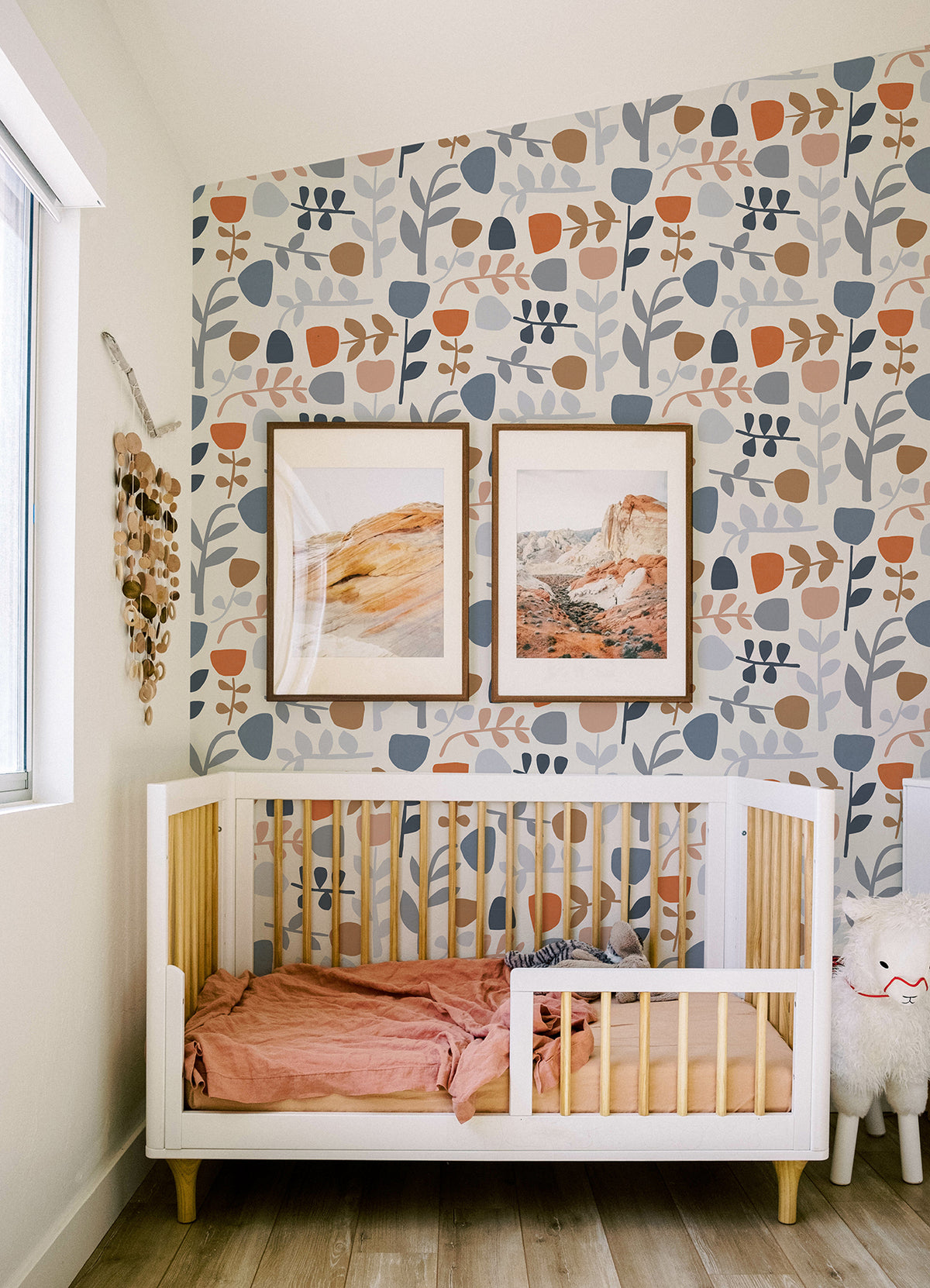 A modern nursery featuring a white crib, pink bedding, a plush toy, wooden wall decor, and two framed landscape prints. The walls are adorned with York Wallcoverings’ Juni Abstract Tulips Wallpaper in blue, peach, and beige tones, while natural light from a large window brightens the space.