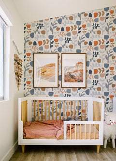 A modern nursery featuring a white crib, pink bedding, a plush toy, wooden wall decor, and two framed landscape prints. The walls are adorned with York Wallcoverings’ Juni Abstract Tulips Wallpaper in blue, peach, and beige tones, while natural light from a large window brightens the space.