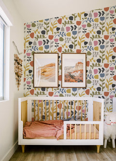 A modern nursery featuring a white crib, pink bedding, Juni Abstract Tulips Wallpaper by York Wallcoverings, framed landscape art, a window, and a plush sheep toy on a wooden floor.