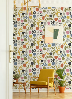A mid-century modern room featuring a yellow armchair, potted plants, and abstract wall art. The wall is adorned with York Wallcoverings’ Juni Abstract Tulips Wallpaper in vibrant shades of pink, yellow, blue, and green. A gold chandelier hangs from the ceiling.