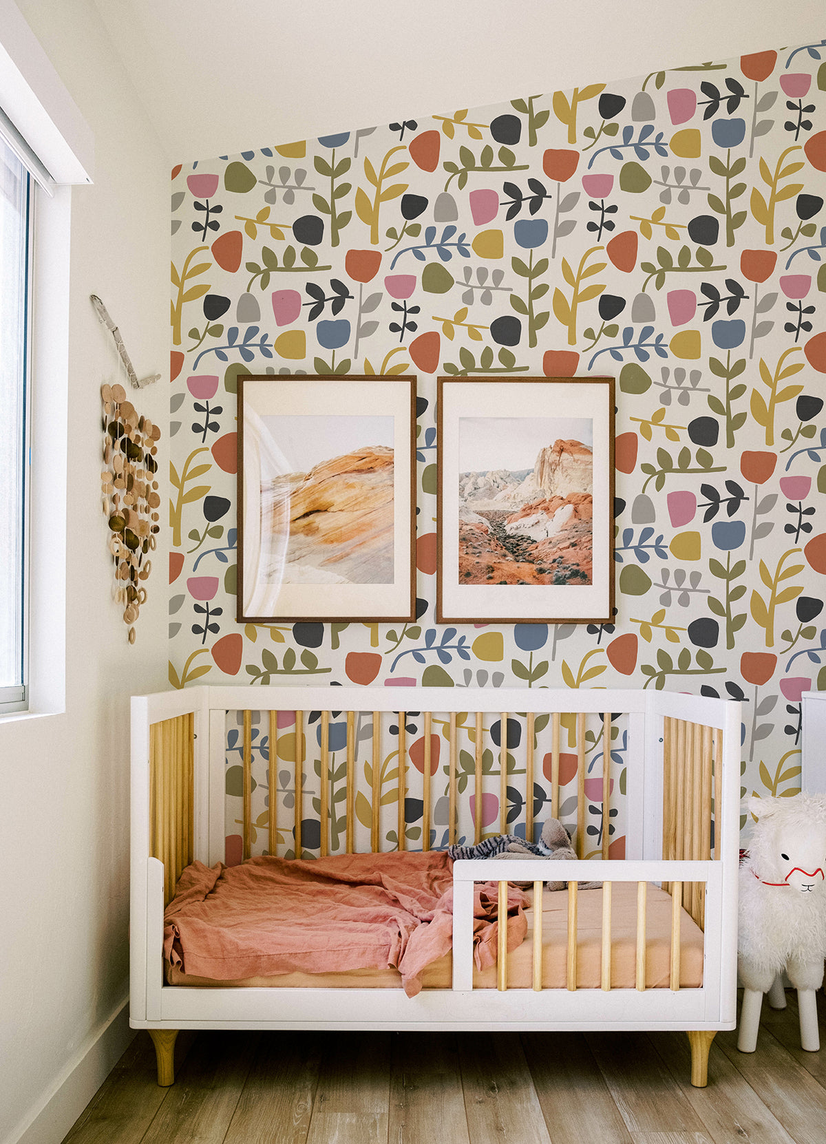 A modern nursery featuring a white crib with pink bedding, Juni Abstract Tulips Wallpaper by York Wallcoverings, two framed landscape prints, and a plush toy beside the crib on a wooden floor.