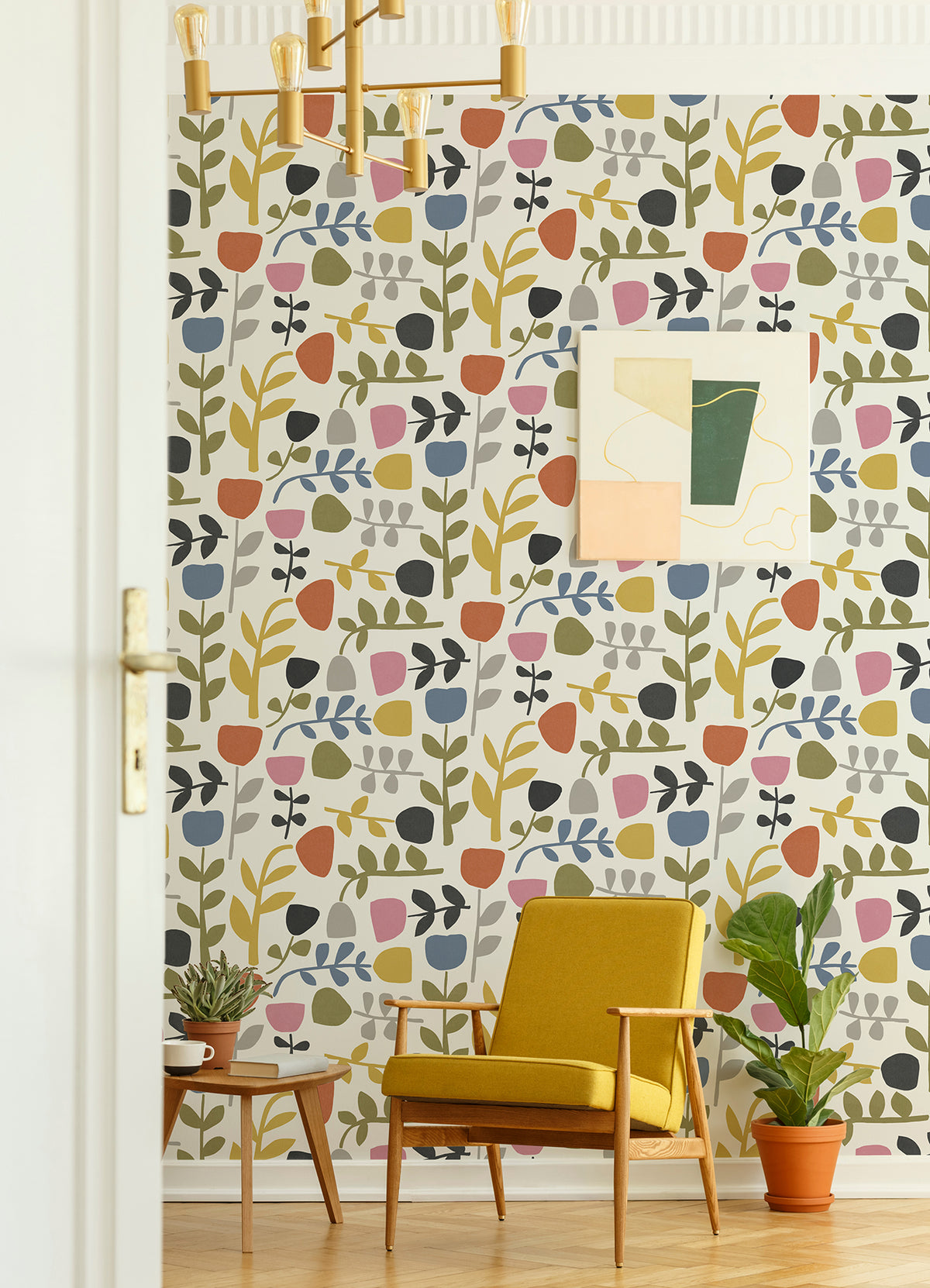 A mid-century modern room with a yellow armchair, plants, abstract wall art, and York Wallcoverings’ Juni Abstract Tulips Wallpaper featuring a colorful, retro floral pattern in green, pink, yellow, and blue tones. A brass chandelier hangs from the ceiling.