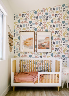 A cozy nursery with a white crib, pink bedding, Juni Abstract Tulips Wallpaper by York Wallcoverings, two framed landscape art pieces, and a plush sheep toy on the floor. Sunlight streams in from a window on the left.