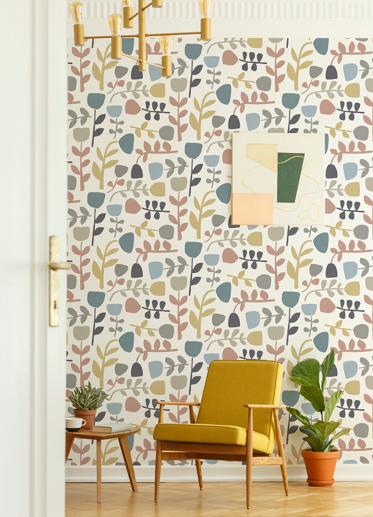 A mustard yellow armchair sits by a small table with potted plants, in front of a wall adorned with Juni Abstract Tulips Wallpaper by York Wallcoverings and abstract art. A modern gold chandelier hangs from the ceiling.