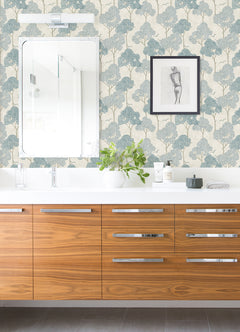 Modern bathroom featuring a wood vanity, white countertop, mirror, and plant. The walls are adorned with Lykke Textured Tree Wallpaper by York Wallcoverings in a blue and white pattern. A framed sketch hangs above various toiletries on the counter.