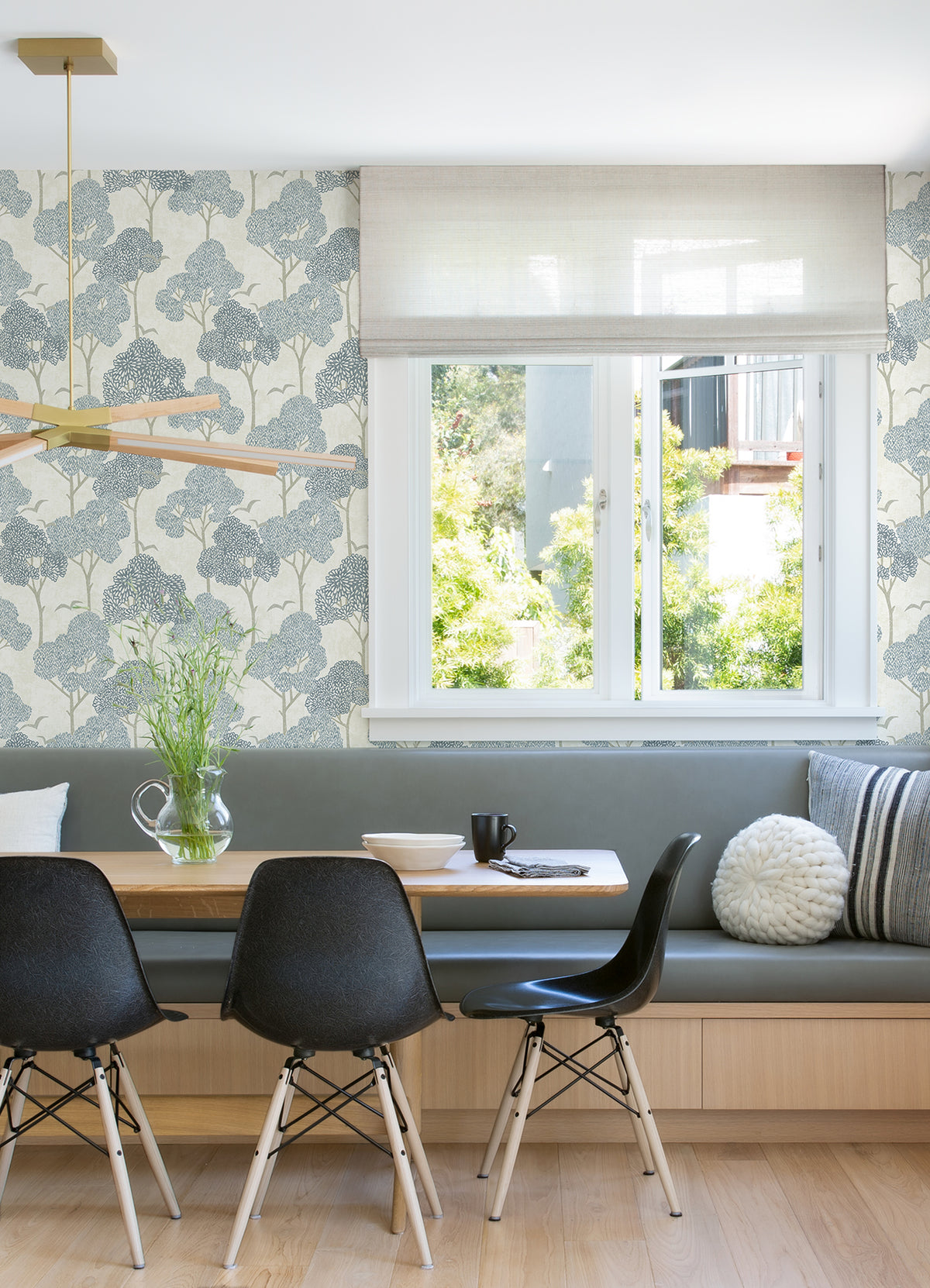 Modern dining nook with a built-in bench, three black chairs, and a wooden table adorned with a glass vase. The walls feature Lykke Textured Tree Wallpaper by York Wallcoverings, and the space is brightened by natural light streaming through a large window onto light wood flooring for a cozy atmosphere.
