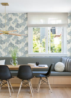 A dining nook with black chairs, a wooden table, a built-in bench with gray cushions, and a window features York Wallcoverings’ Lykke Textured Tree Wallpaper on the wall. A vase of flowers sits on the table.